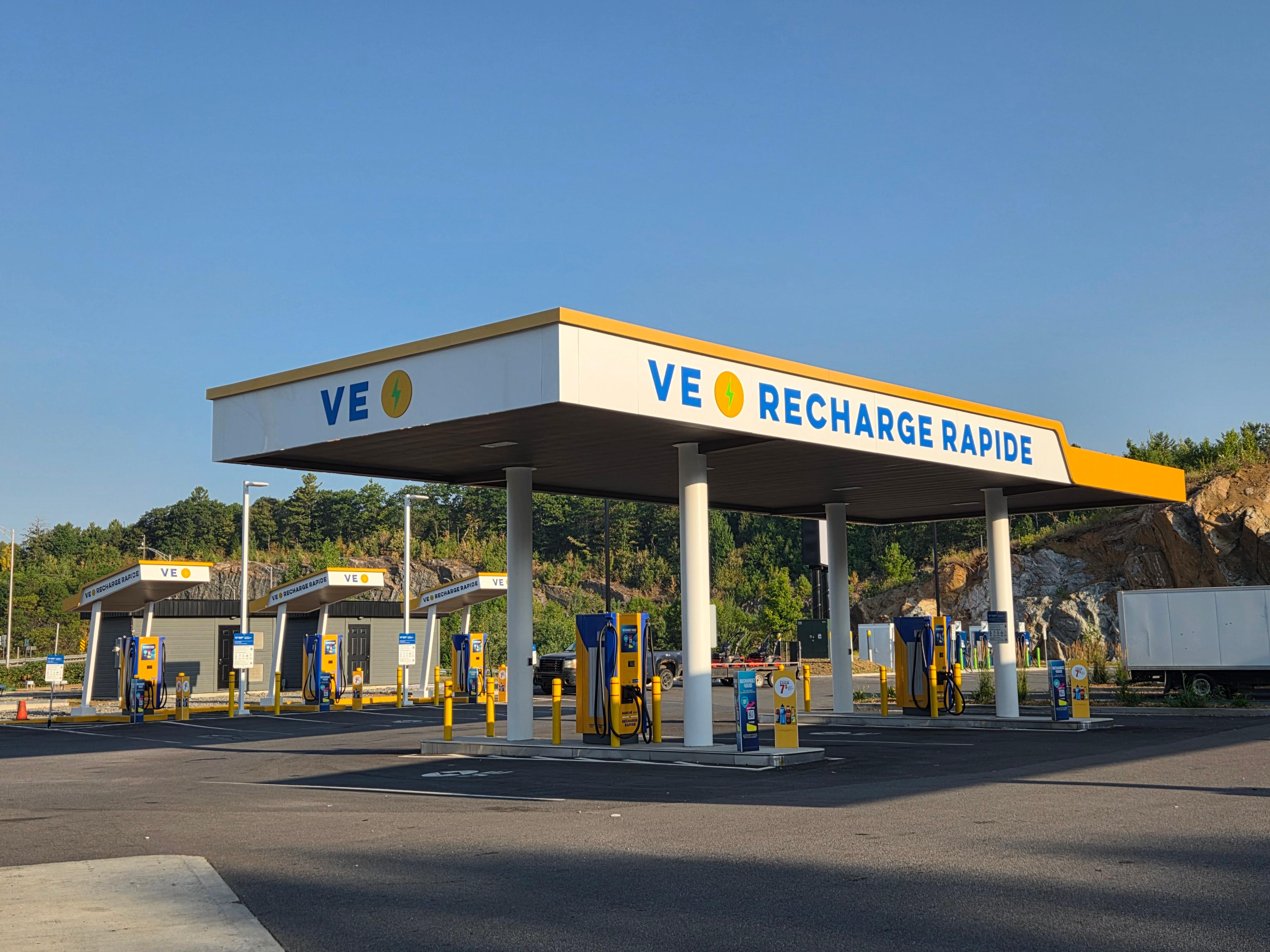La photo montre une station VE Recharge Rapide avec plusieurs bornes de recharge jaunes et bleues sous un grand auvent blanc. L’enseigne affiche clairement « VE Recharge Rapide » en lettres bleues avec une icône d’éclair jaune. Le site est propre et spacieux, avec plusieurs bornes alignées et un marquage clair au sol.