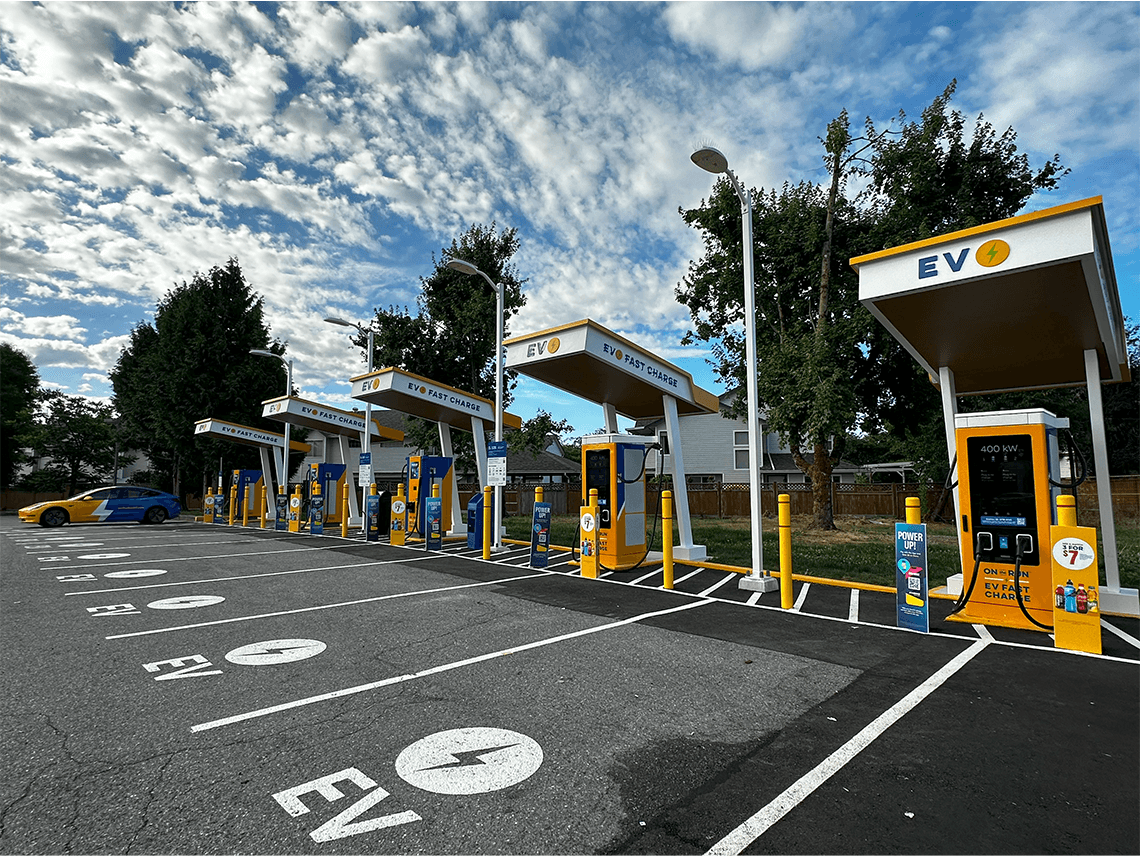 The photo shows an EV Fast Charge station with multiple yellow and blue chargers under white canopies. The parking spots are clearly marked with EV symbols. A blue and yellow EV with a large white lightning bolt graphic is parked at one of the stalls.