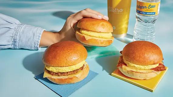 Hand holding a breakfast sandwich with other sandwiches in the foreground.